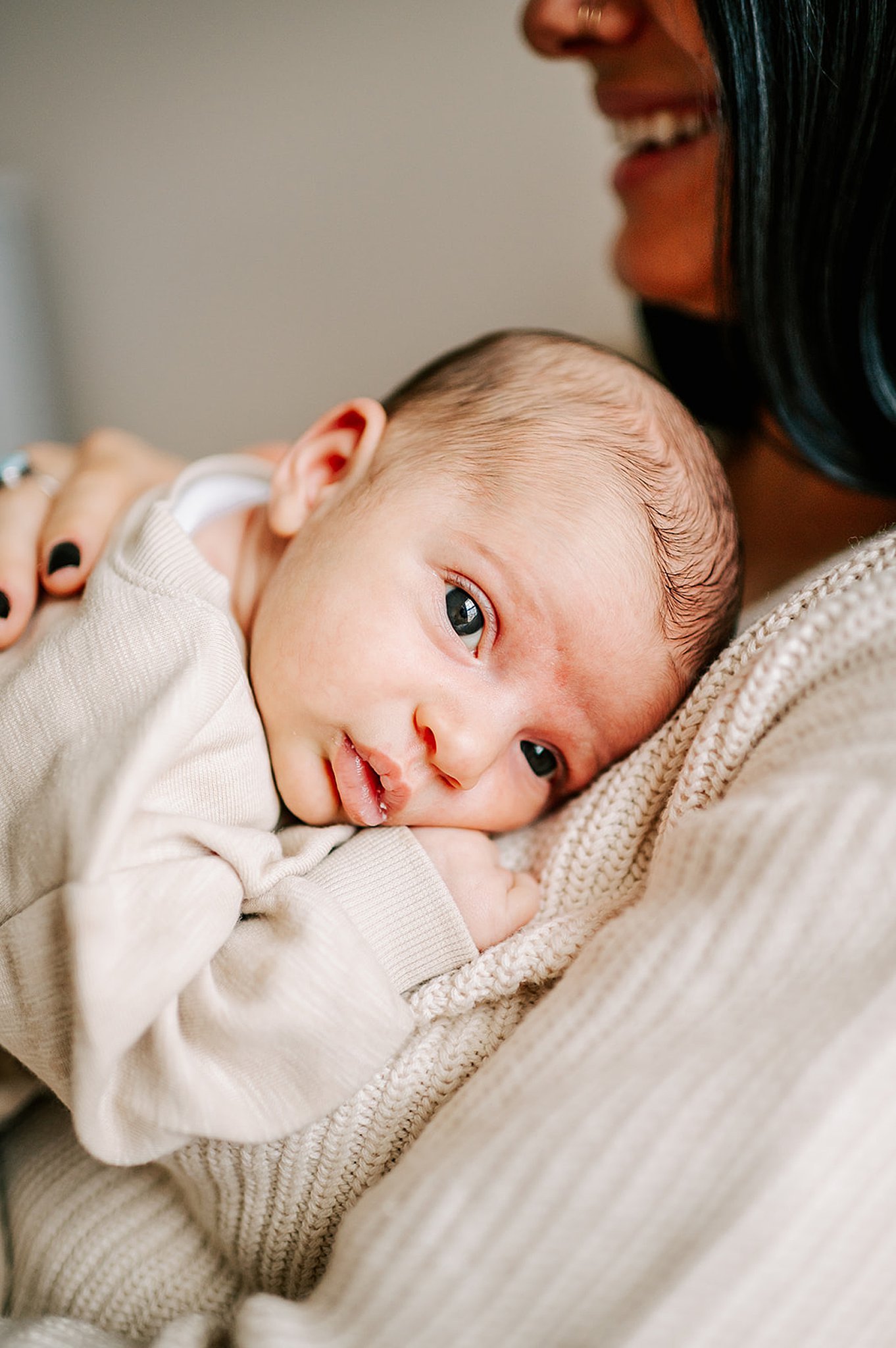 A newborn baby lays on mom's chest with eyes open in a tan sweater