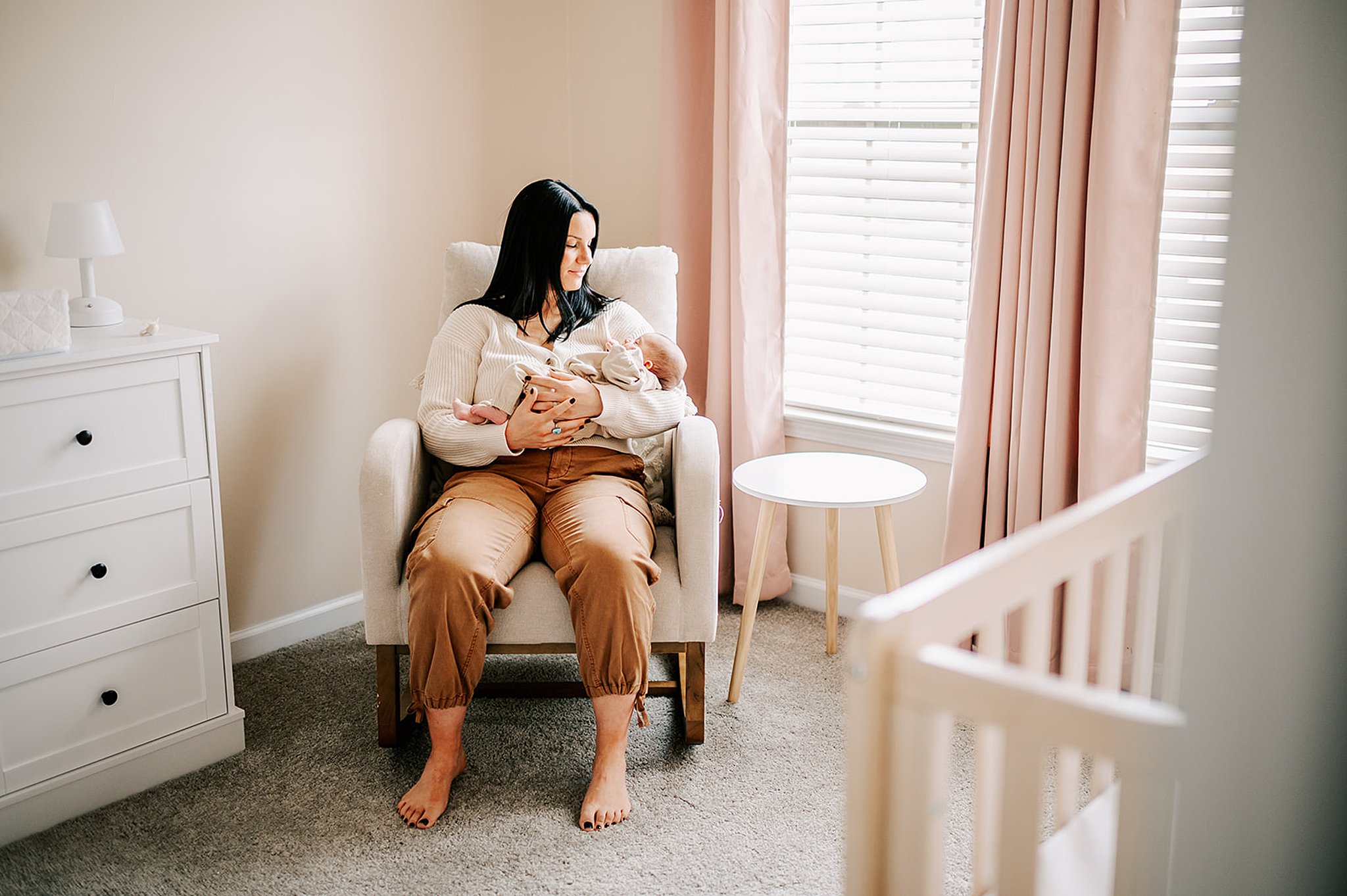 A smiling new mom sits in a nursing chair in a nursery under a window cradling her newborn baby in her arms after visiting wynnies boutique