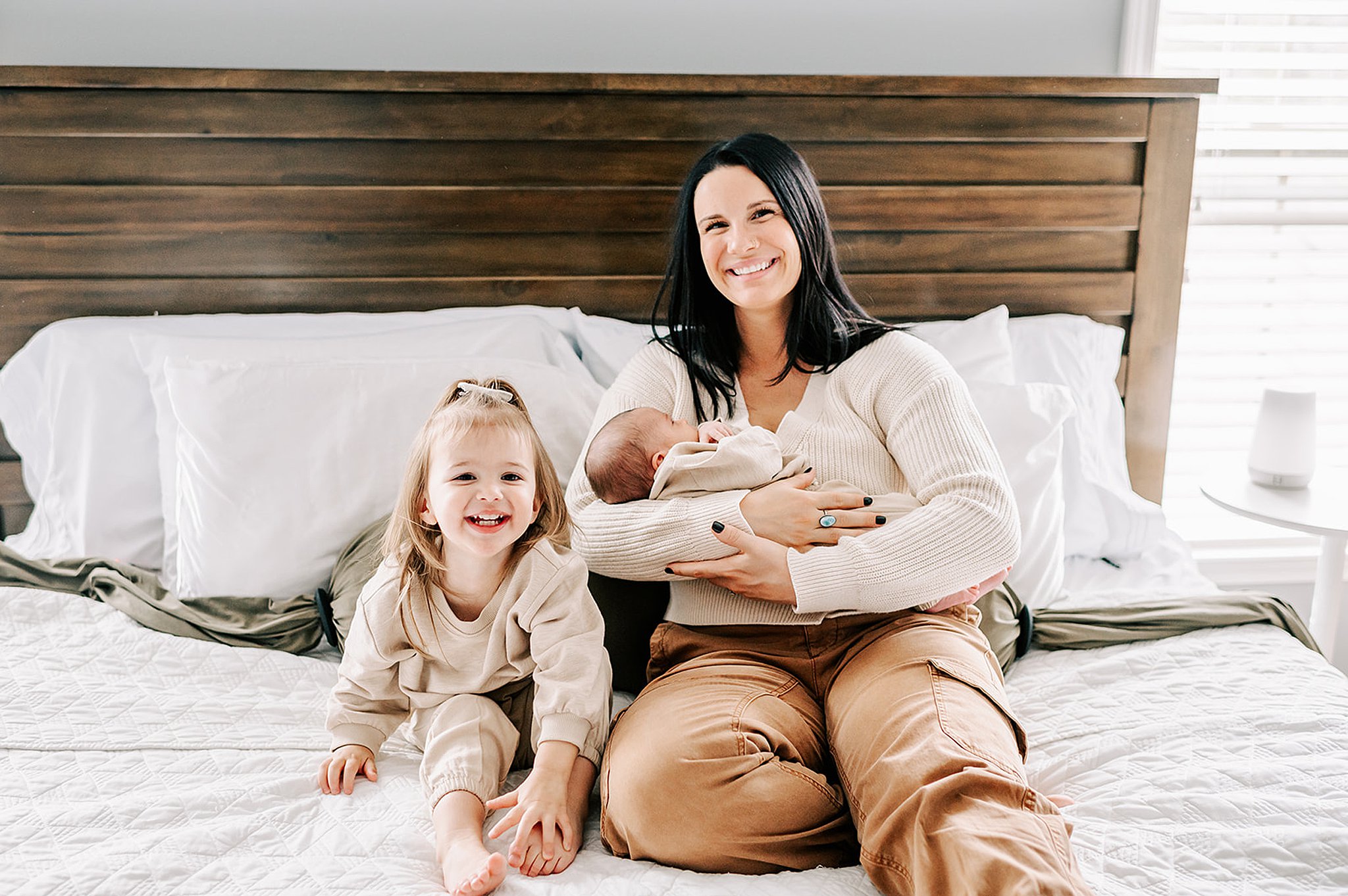 A toddler girl giggles while sitting on a bed with mom cradling her newborn baby sister after visiting wynnies boutique