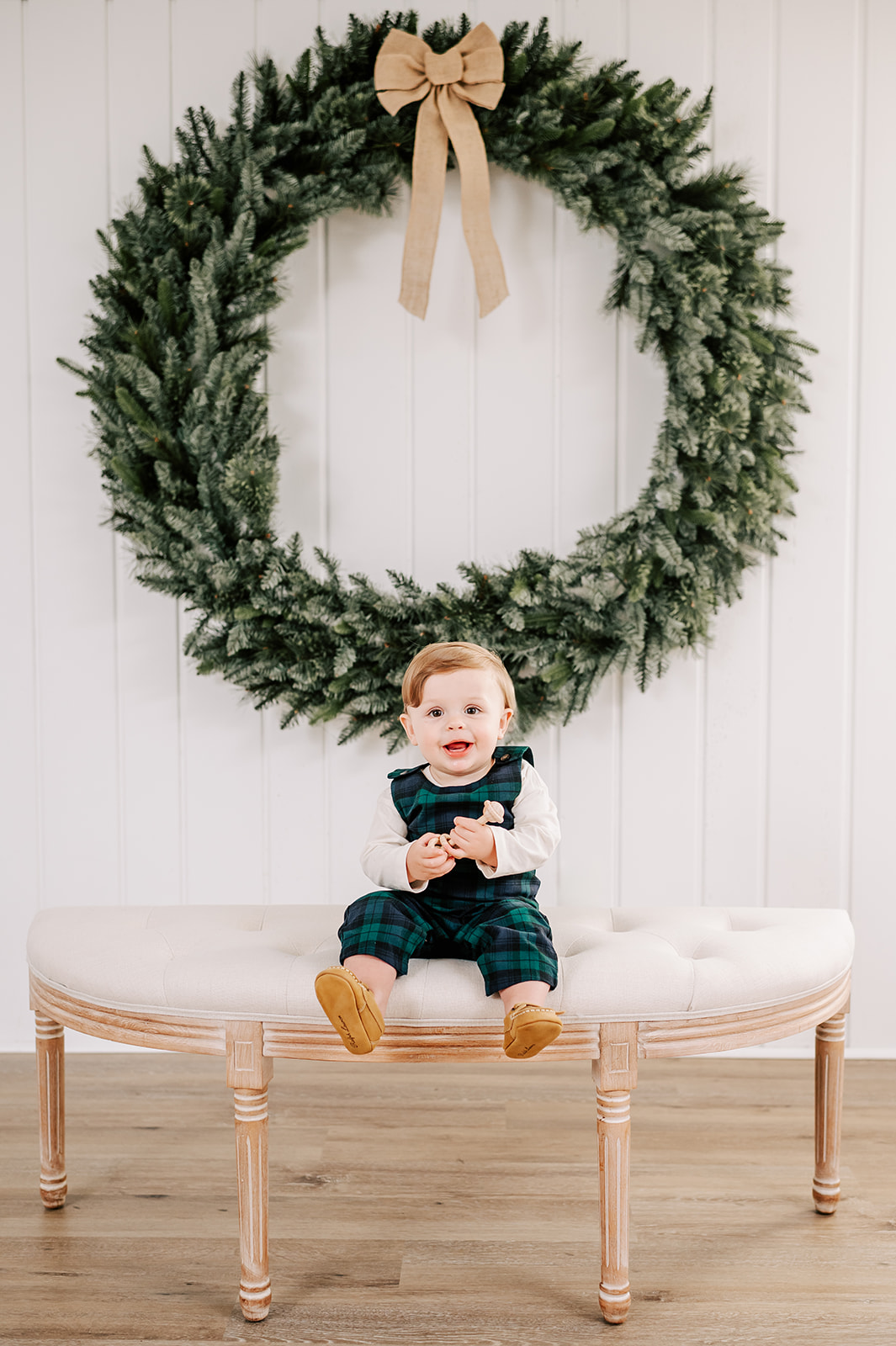 A smiling baby sits on a bench in a studio with a christmas wreath hanging behind him