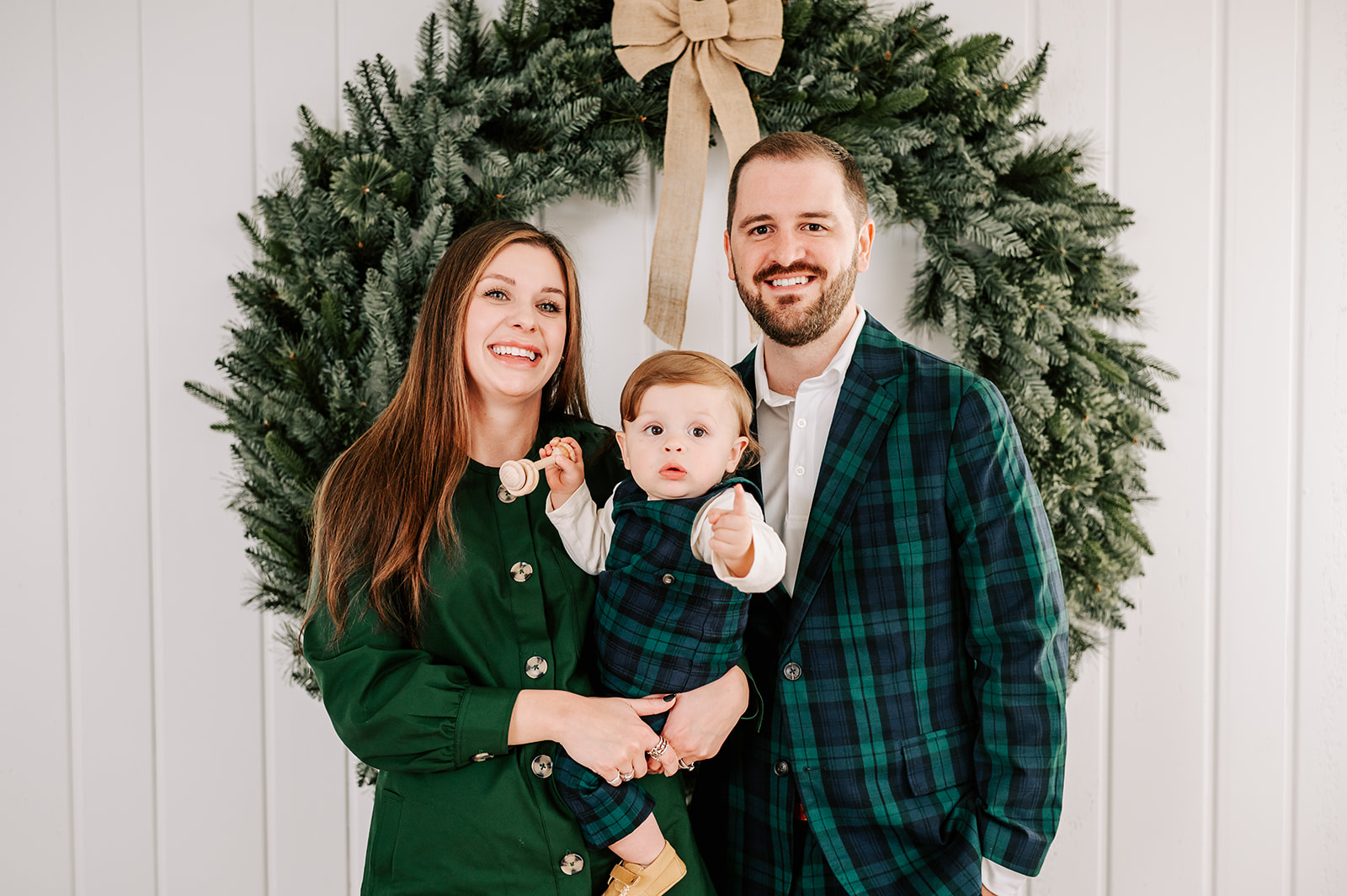 A happy family of 3 stand in front of a christmas wreath in a studio with the baby pointing and holding a wooden toy before visiting Santa in WInston-Salem