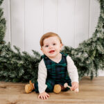 A baby in green plaid overalls sits on the floor playing with a wooden toy in front of a Christmas wreath before visiting Santa in WInston-Salem