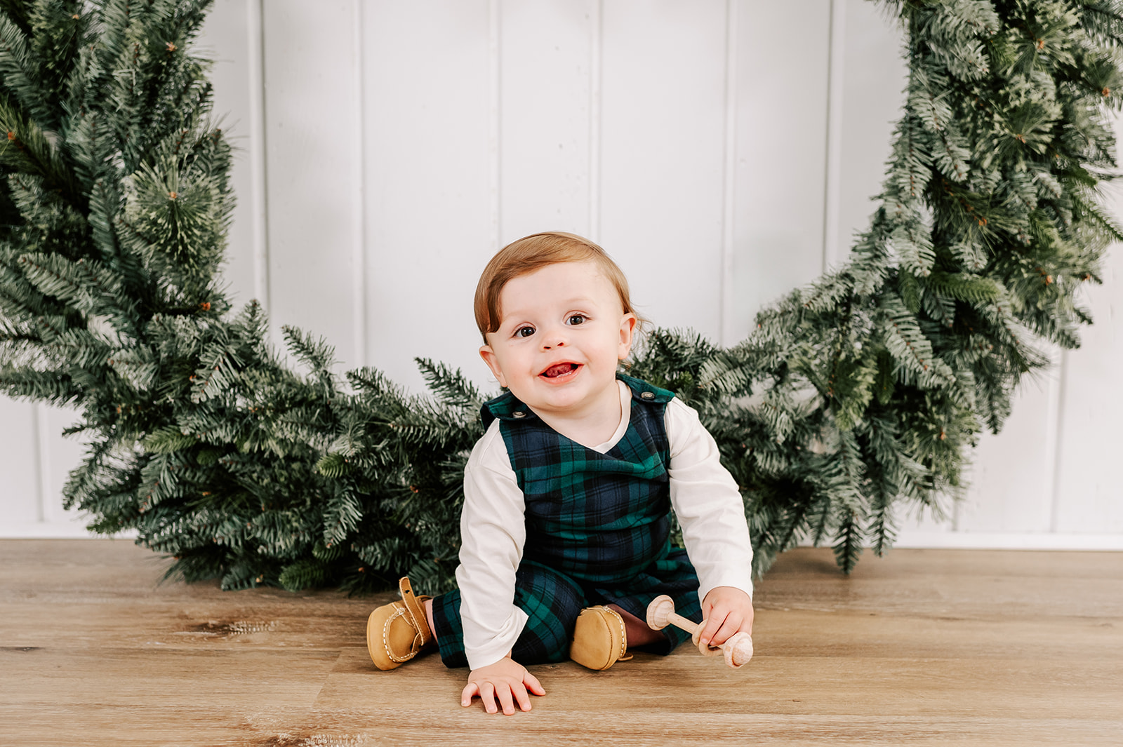 A baby in green plaid overalls sits on the floor playing with a wooden toy in front of a Christmas wreath before visiting Santa in WInston-Salem