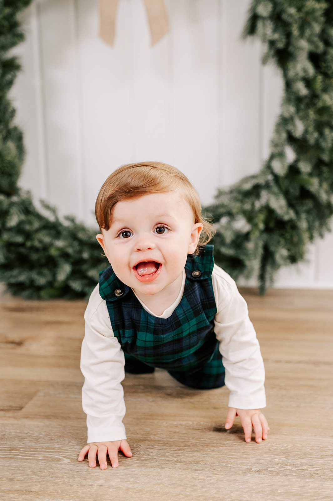 A baby in plaid overalls crawls on the floor of a studio in front of a christmas wreath before visiting Santa in WInston-Salem