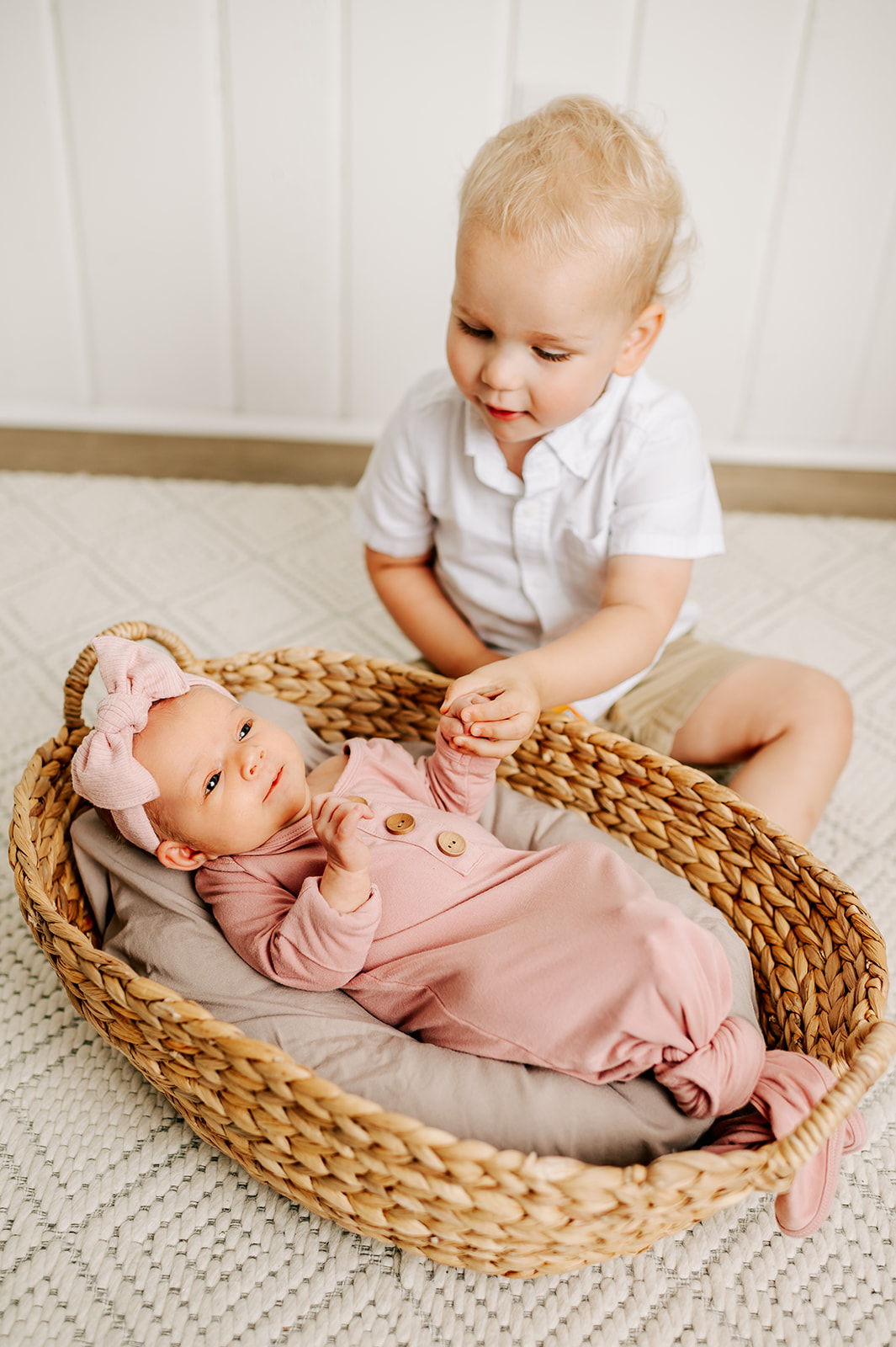 A newborn baby girl in a pink swaddle and matching bow holds hands with toddler older brother while laying in a woven basket in a studio