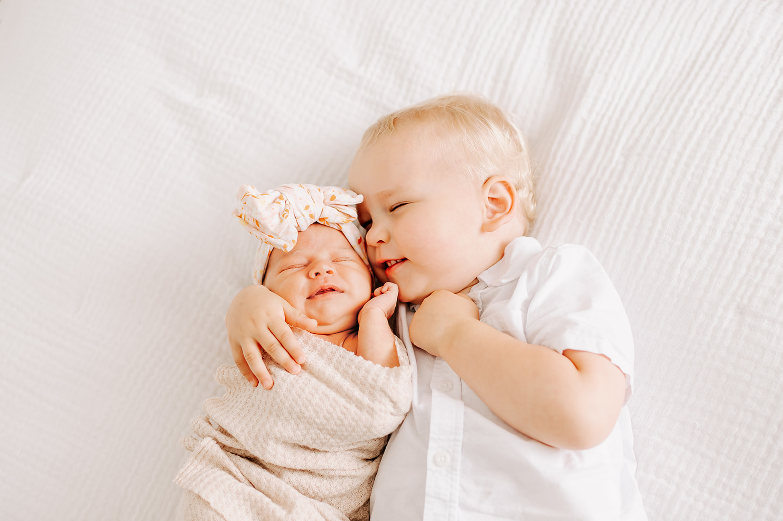 A toddler boy in a white shirt snuggles cheek to cheek with his newborn baby sister on a white bed before visiting Carowinds Winterfest