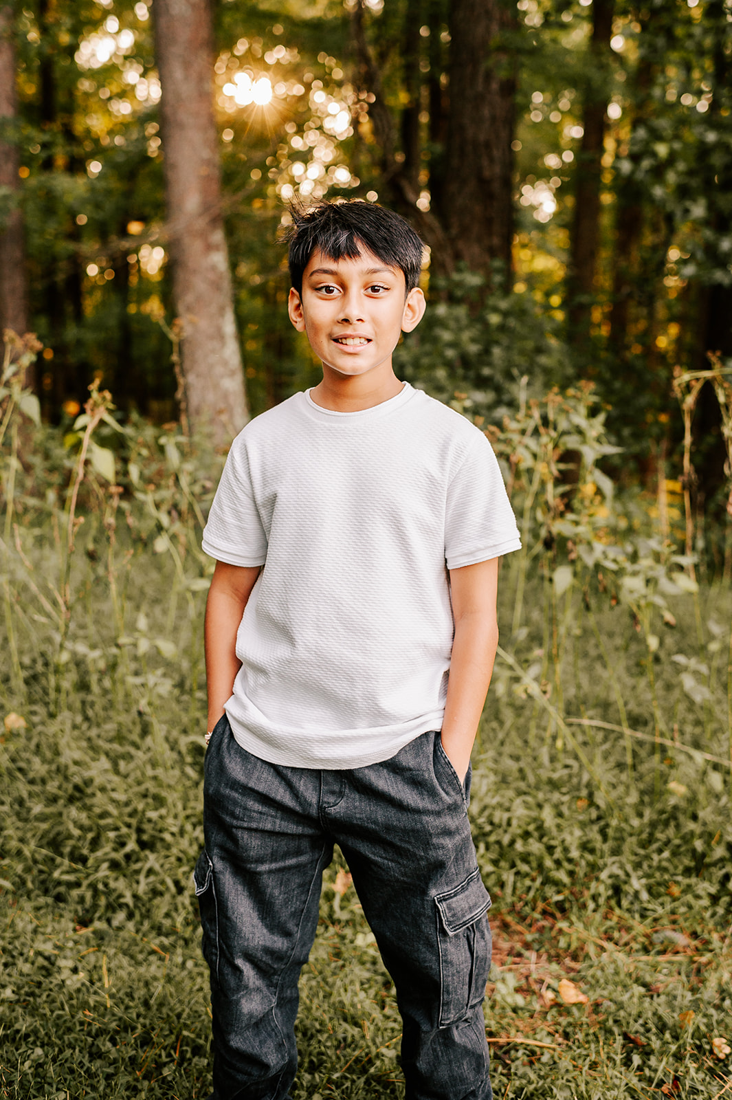 A young boy in a white shirt and jeans stands on the edge of a forest at sunset with hands in his pockets