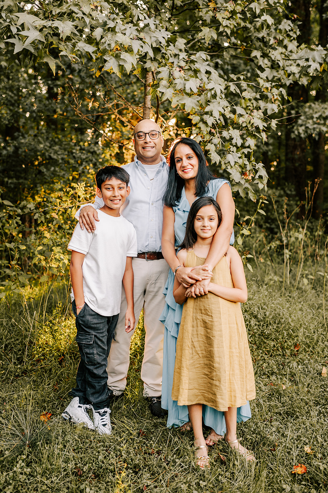 A smiling mom and dad hug and hold their young son and daughter on the edge of a forest at sunset before visiting holiday events in Winston-Salem, NC