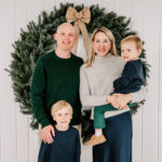 Happy mom and dad stand in a studio in front of a wreath with their two toddler sons before visiting the Winston-Salem Holiday Parade