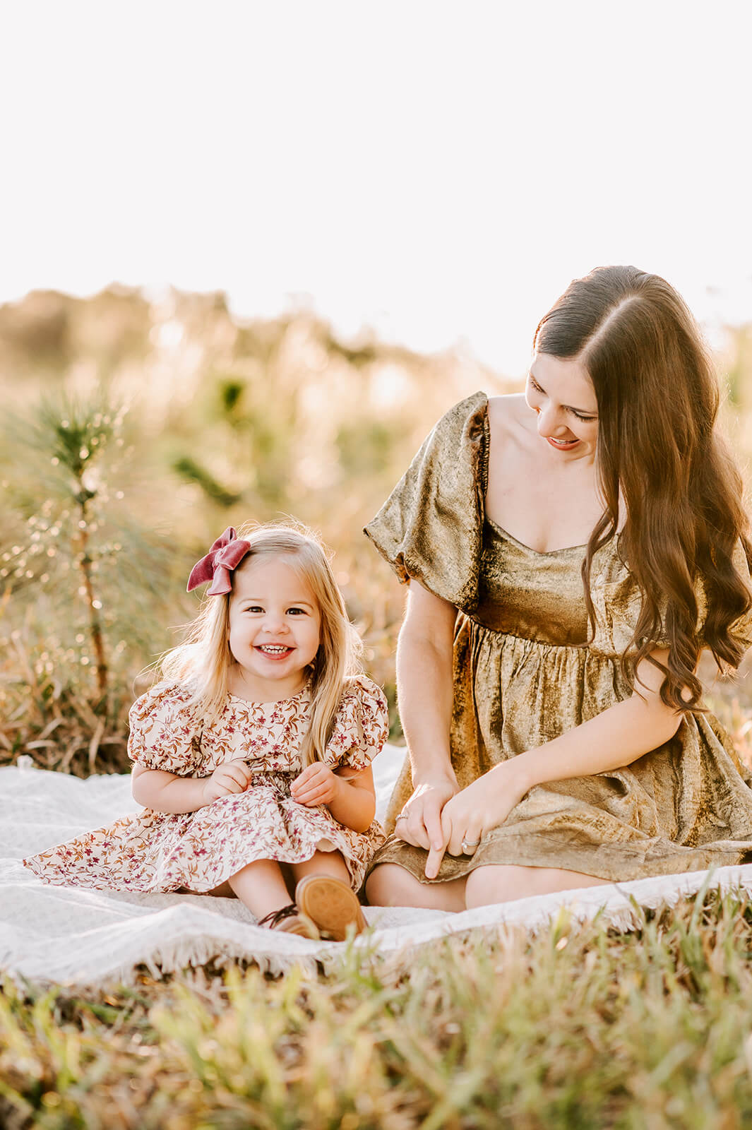 A toddler girl in a floral print dress and pink bow smiles big while sitting in a blanket with mom after visiting bella and james