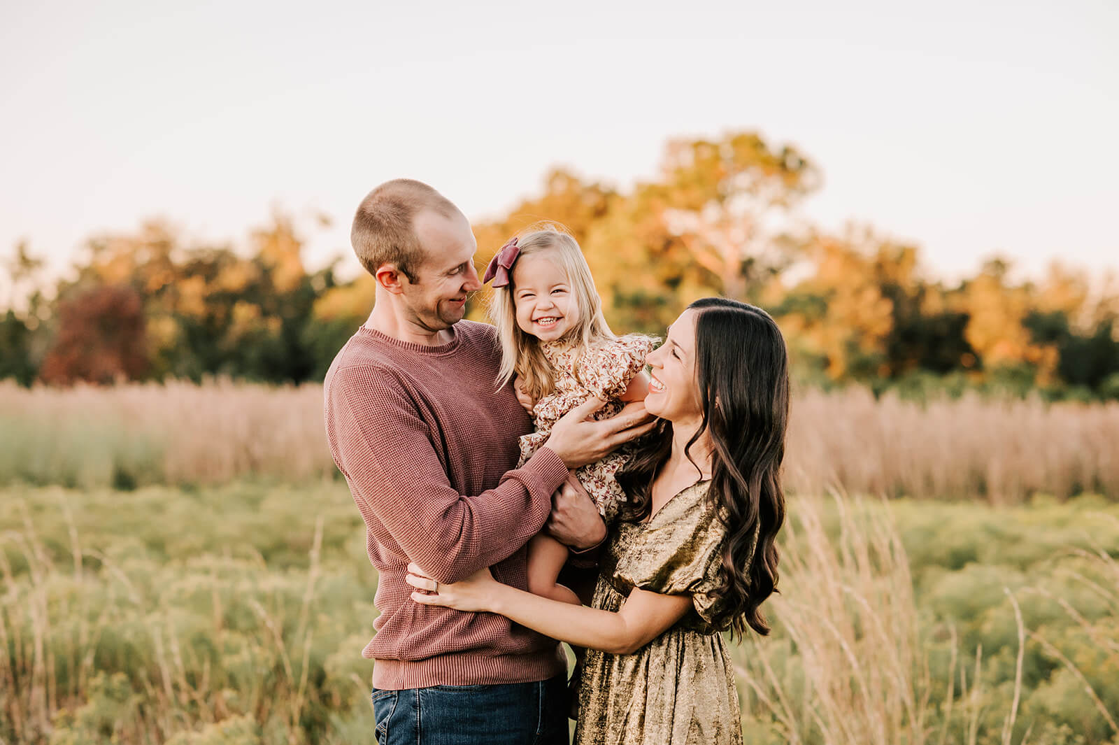A happy toddler girl in a dress and pink bow sits in mom and dad's arms while exploring a park at sunset with tall grass after visiting bella and james