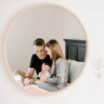 A peak at mom and dad snuggling and cradling their newborn baby on a bed through a mirror after visiting children's boutiques in winston salem