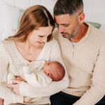 Happy new parents in cream sweaters snuggle their newborn baby in mom's arms while sitting on a bed before visiting the nest schools