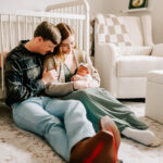 A happy couple snuggles on the floor of their nursery while cradling their sleeping newborn daughter