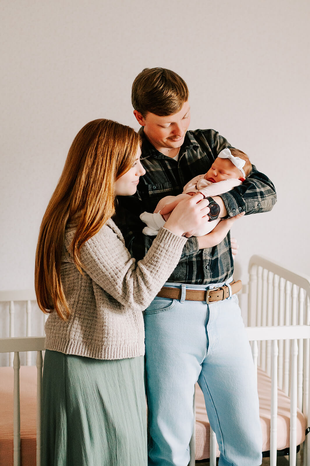 A happy couple snuggles their sleeping newborn daughter in dad's arms while standing in the nursery before finding baby music classes in winston-salem