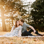 A happy expecting couple snuggles as he kisses her cheek on a picnic blanket in a park lawn at sunset