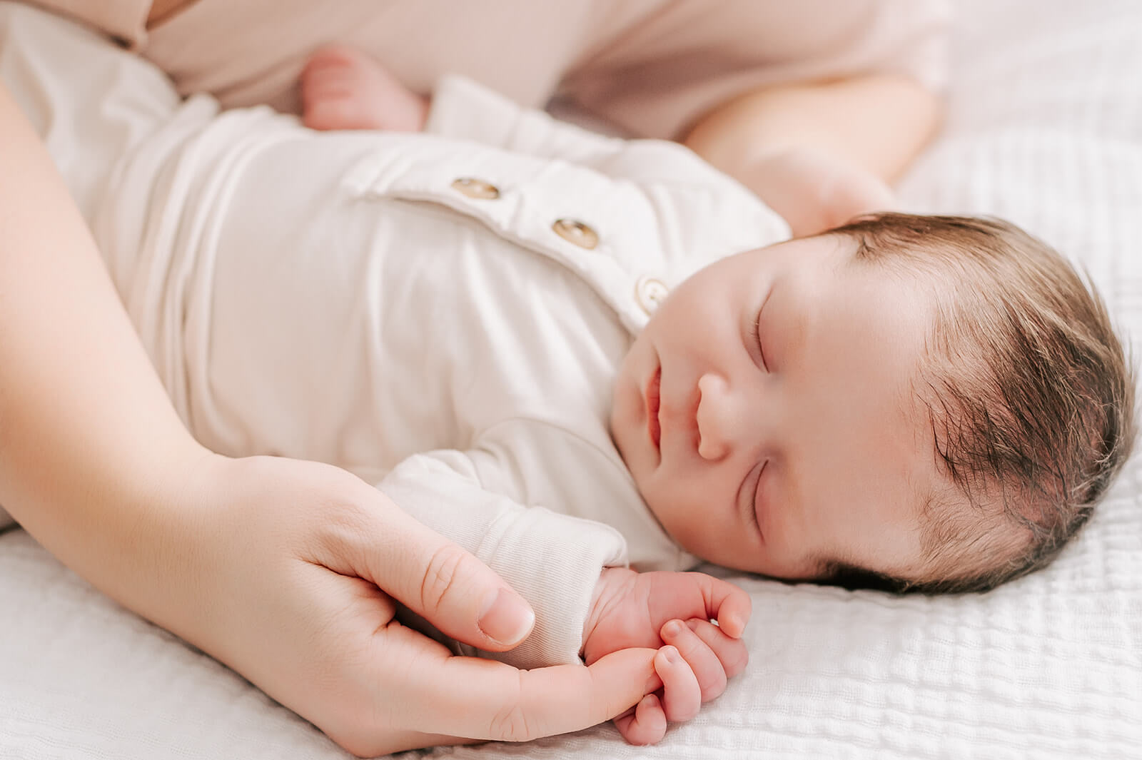 A newborn baby sleeps in a white onesie on a bed while holding mom's finger