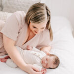 A newborn baby in a white onesie sleeps on a bed in a studio while holding mom's finger as she snuggles before exploring baby swim lessons in winston salem