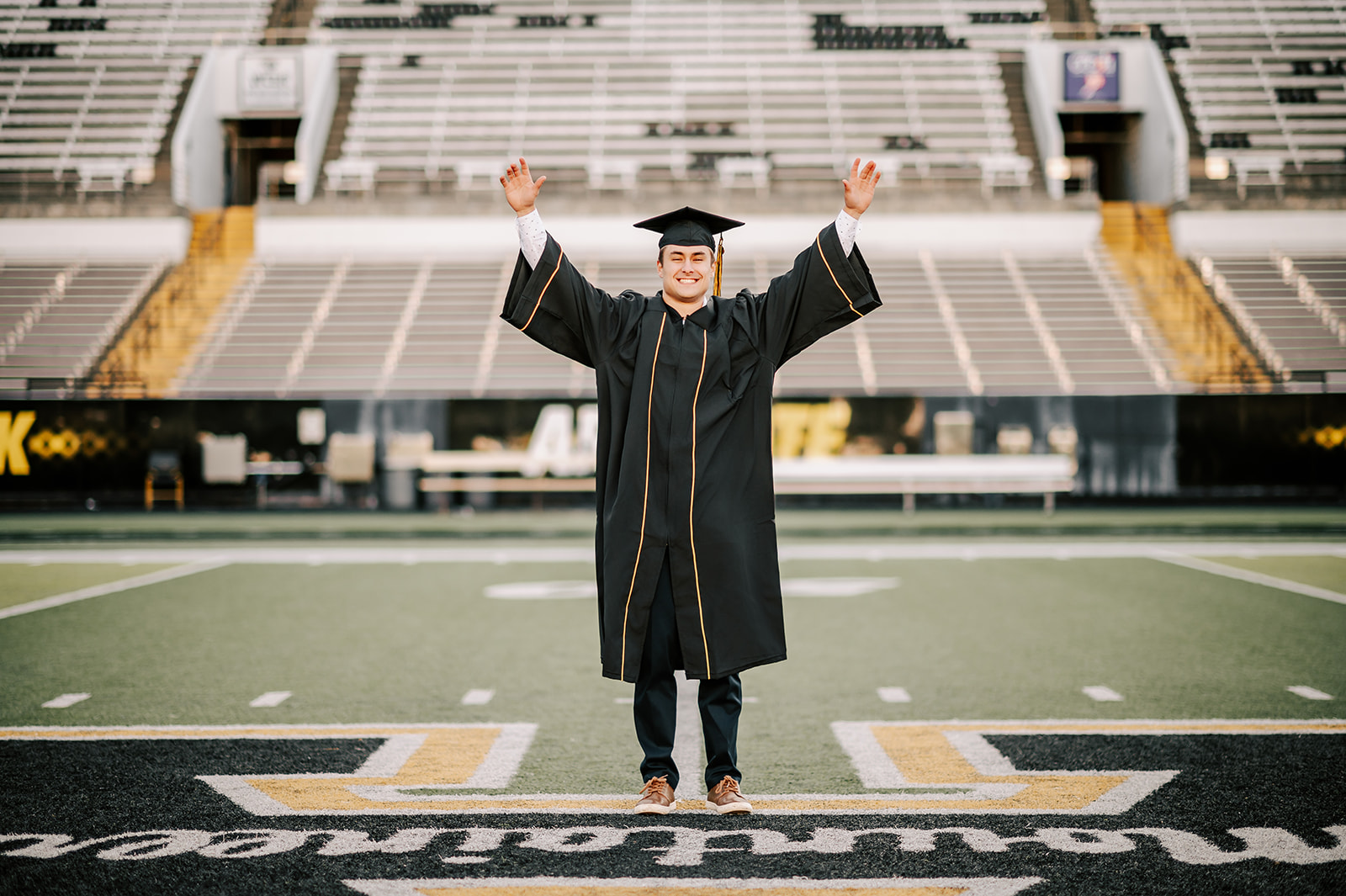 A happy grad in cap and gown lifts his hands while standing in the center of the football field