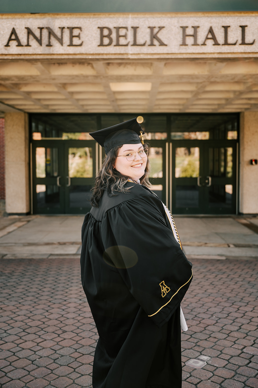 A happy graduate smiles over her shoulder in her cap and gown in front of one of the halls' entrances