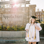 A happy grad in her cap stands with the Appalachian State University sign at sunset