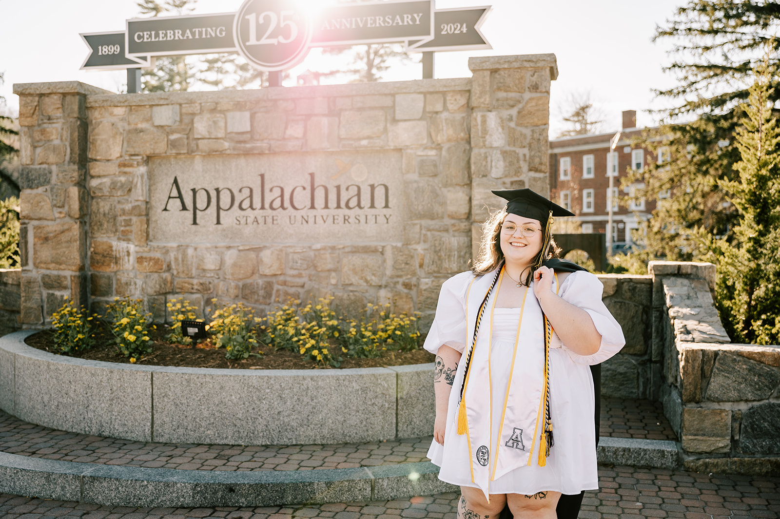 A happy grad in her cap stands with the Appalachian State University sign at sunset