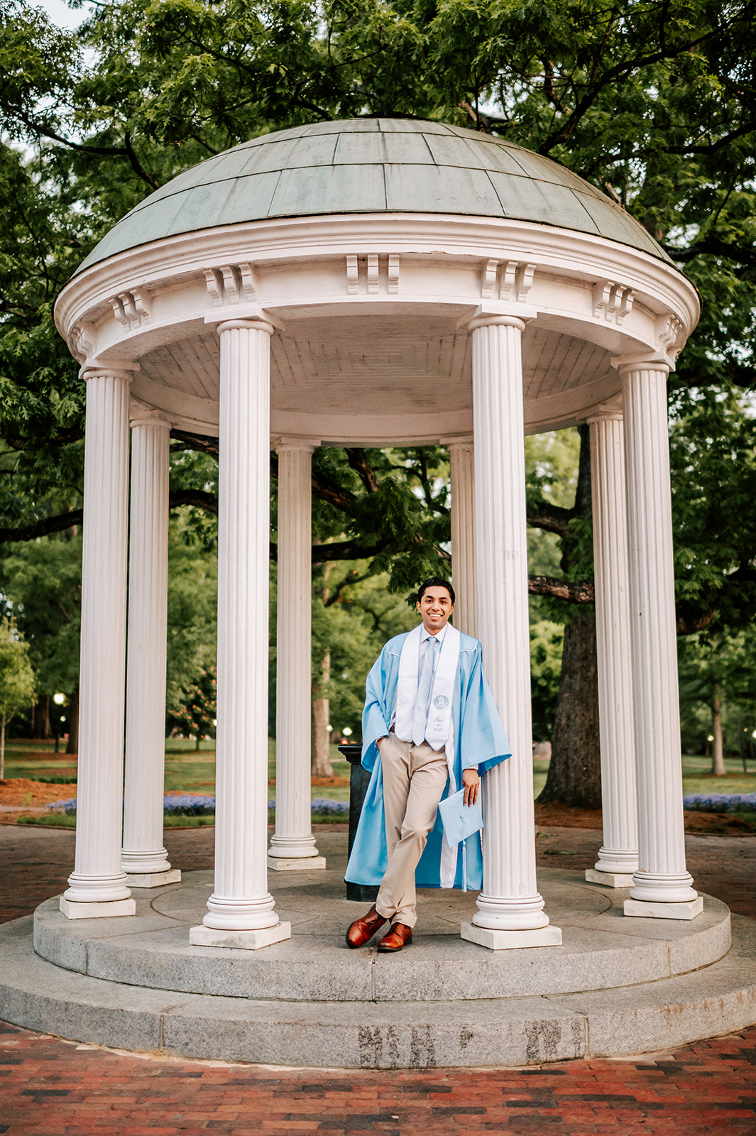 A UNC grad leans on the pillar of the old well at UNC in his gown and khaki pants