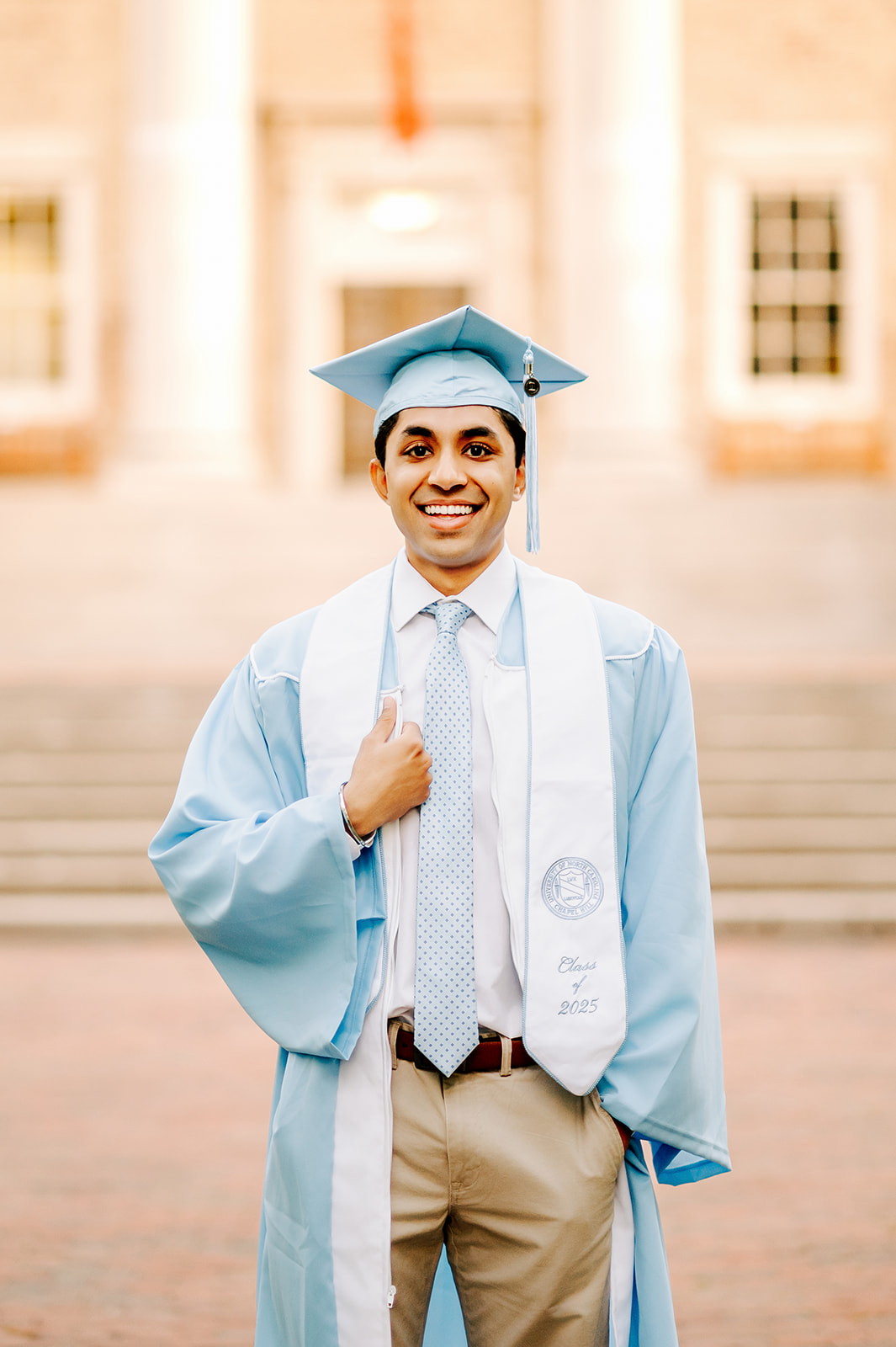 One of the happy UNC-Chapel Hill Graduates stands smiling in his blue cap and gown holding his lapel in front of a building