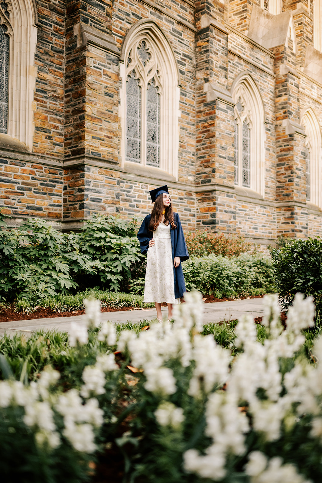 One of the Duke Graduates walks through the chapel gardens in her cap and gown and white dress