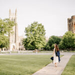 A happy grad walks a sidewalk in the quad towards the chapel while smiling over her shoulder