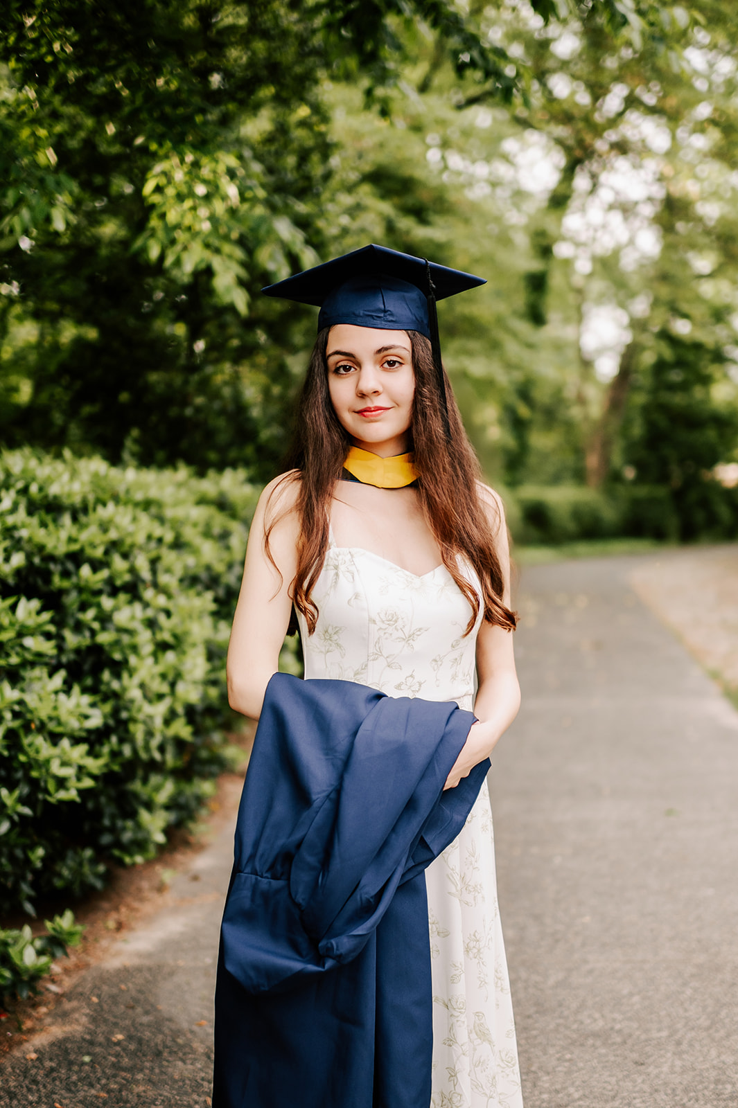One of the beautiful Duke Graduates stands on a sidewalk holding her gown while wearing her cap