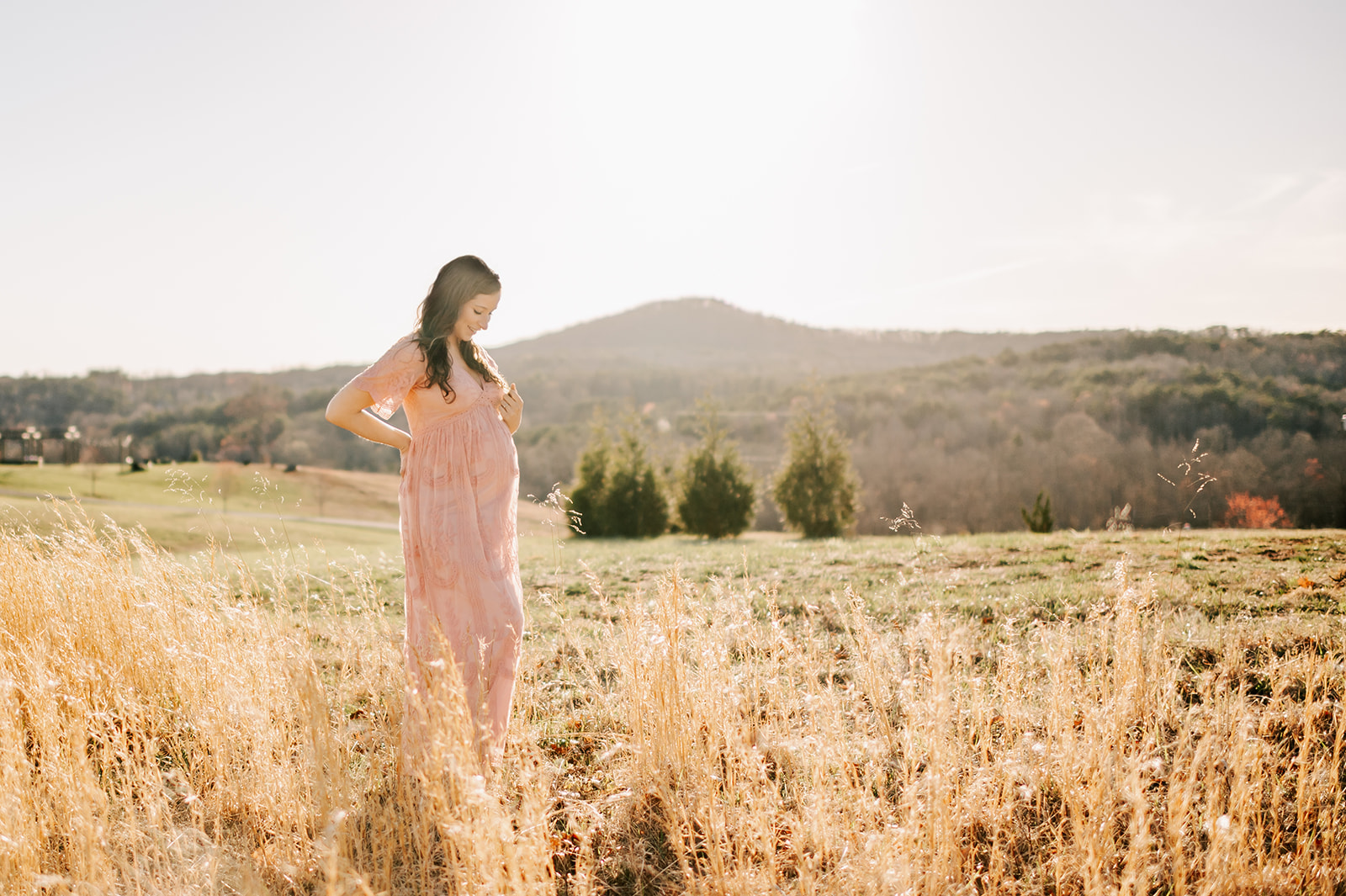 A pregnant woman in a pink maternity gown smiles down to her bump while standing in a pasture at sunset after visiting a birth center in Winston-Salem, NC