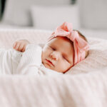 A newborn baby girl sleeps in a basket wearing a white onesie and pink bow while using a diaper service in Winston-Salem