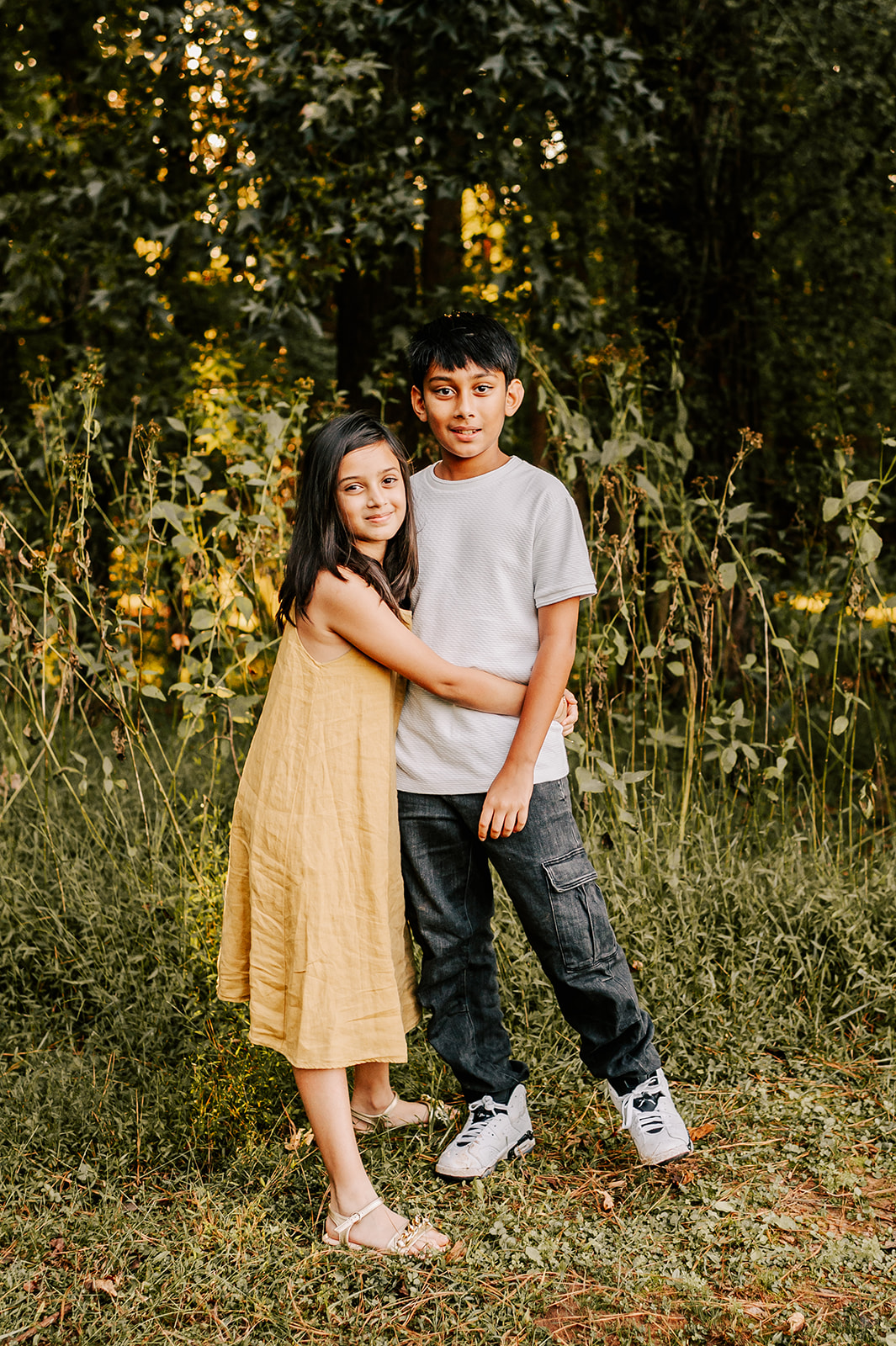 A young girl in a tan dress hugs her older brother as they smile on the edge of the woods