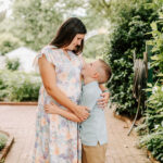 A happy mother in a floral print dress hugs and smiles down to her happy young son in a green shirt in a garden after finding mom groups in Winston-Salem, NC