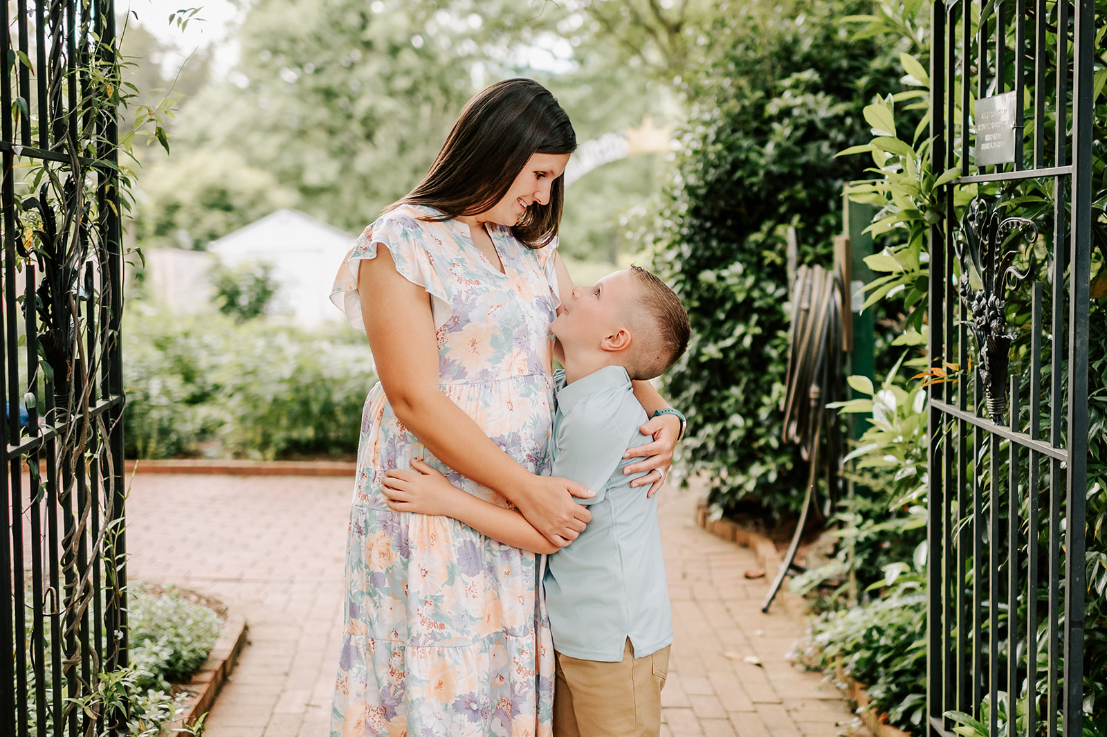 A happy mother in a floral print dress hugs and smiles down to her happy young son in a green shirt in a garden after finding mom groups in Winston-Salem, NC