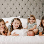 Four toddler sisters lay on a bed behind their newborn baby brother sleeping in a white swaddle