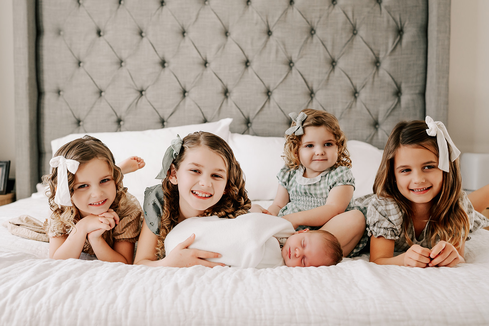 Four toddler sisters lay on a bed behind their newborn baby brother sleeping in a white swaddle