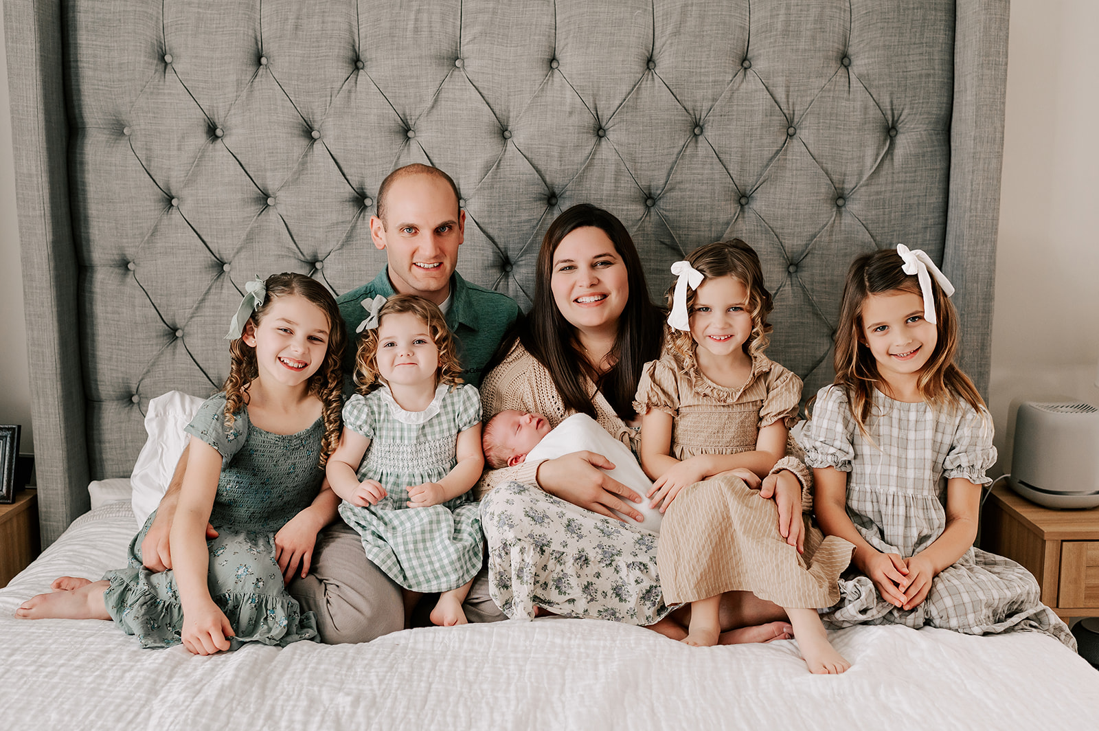 A smiling mom and dad sit on a bed with their four toddler daughters and newborn son in their laps