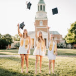 Three Wake Forest grads toss their caps in the air while wearing white dresses and gold sashes at one of the colleges near Winston-Salem