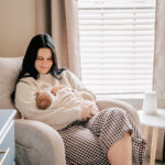 A happy mom sits in a nursing chair cradling her newborn daughter in her lap under a window after meeting lactation consultants in Winston-Salem, NC