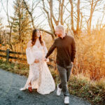 A happy expecting couple walk and laugh while in a mountain trail at sunrise in a long maternity gown and long sleeve shirt