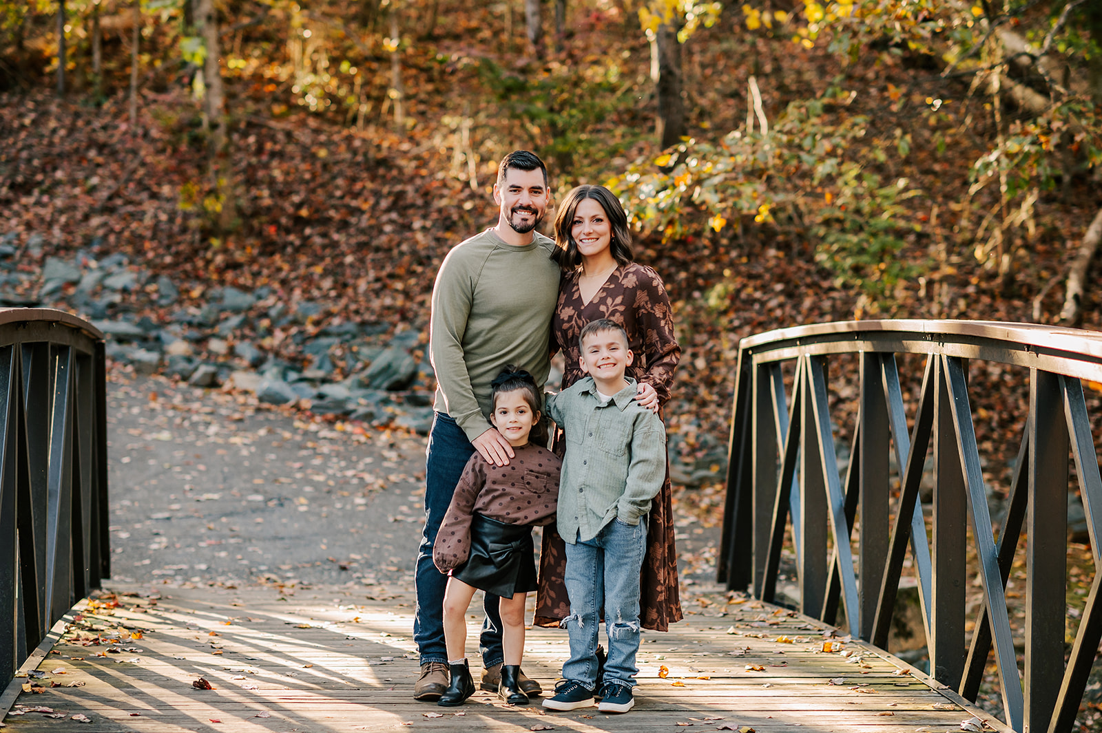 Happy mom and dad stand with their toddler son and daughter in brown and green on a pedestrian park bridge in fall at sunset after meeting a Winston-Salem nanny