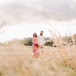 A happy pregnant couple hold hands on a hike in a pink maternity gown and cream shirt in a meadow of tall golden grass