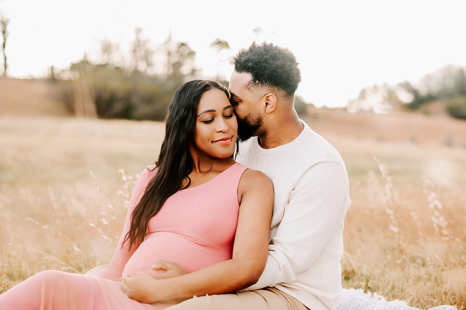 A mom to be smiles while leaning into her partner in a field of tall golden grass at sunset after expliring Mom League