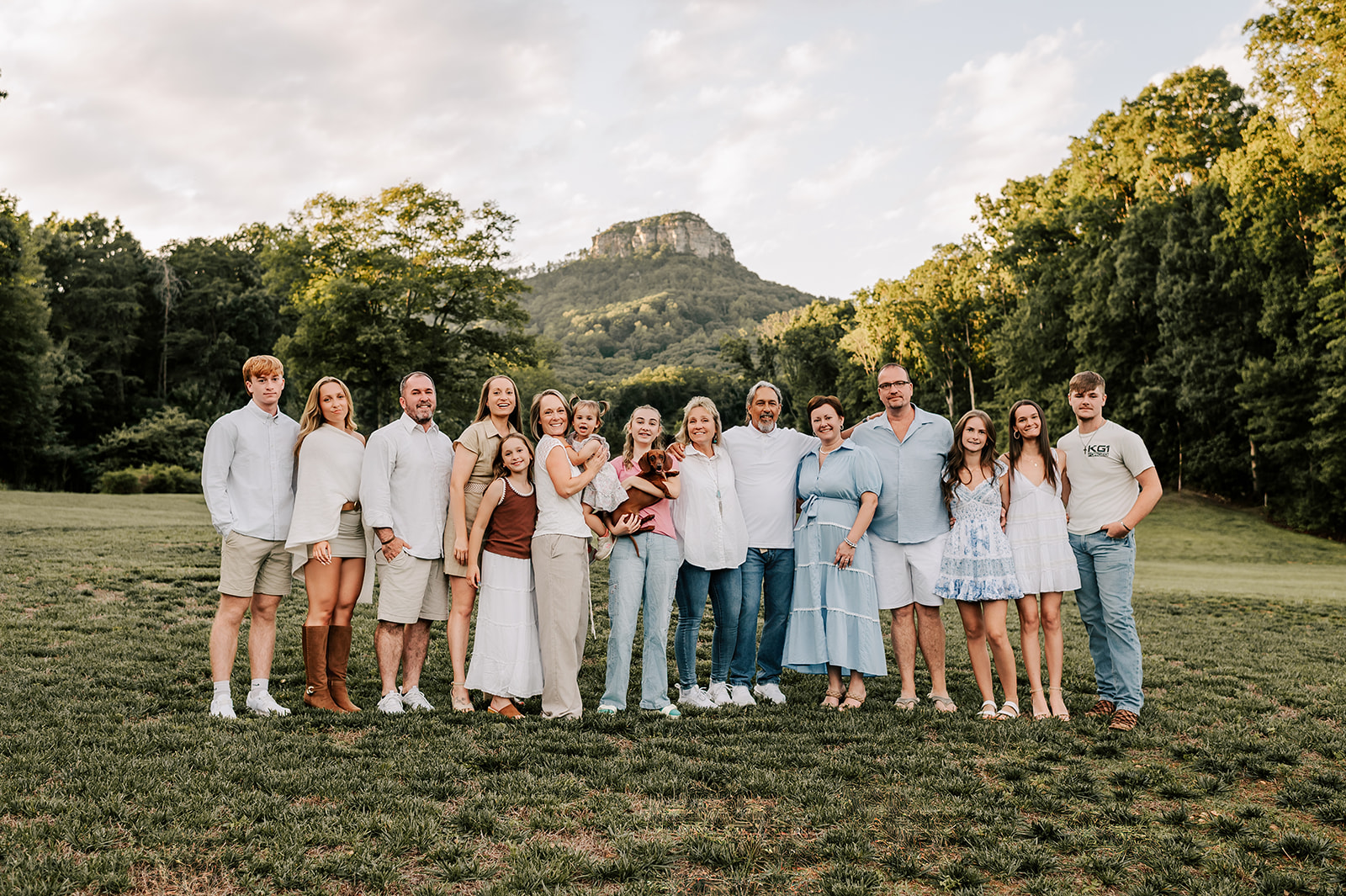 A large extended family stands smiling in a field near pilot mountain at sunset