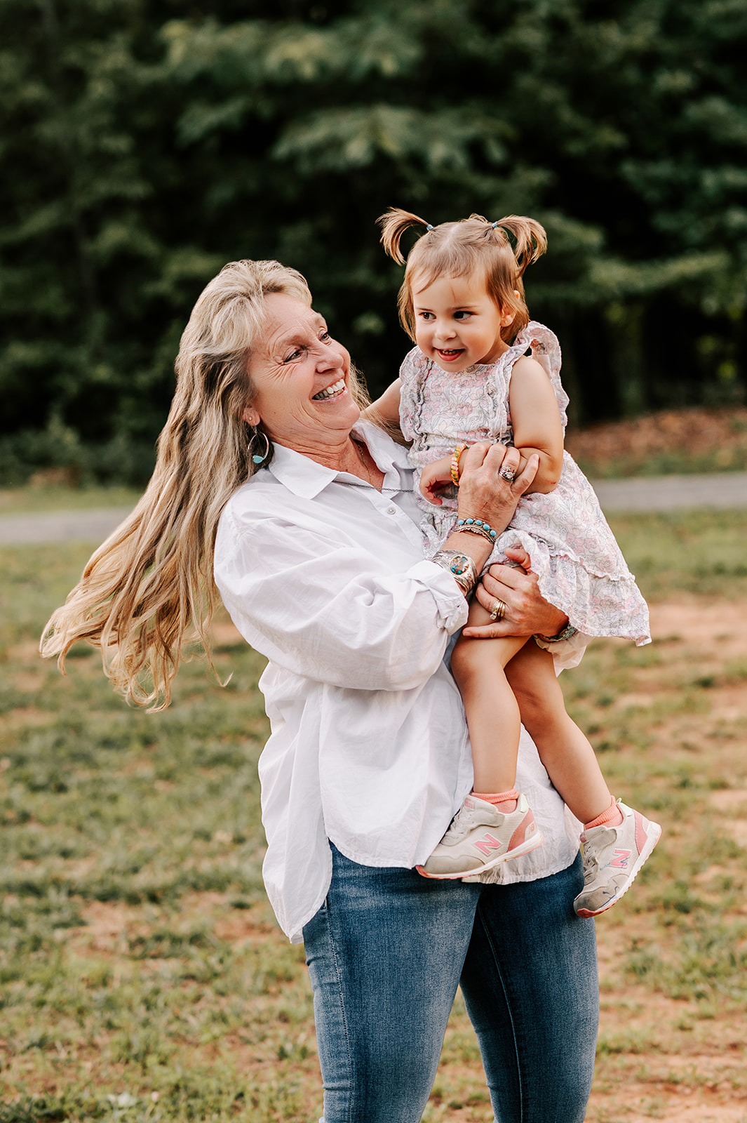 A happy grandma plays with her toddler granddaughter on her hip in a field at sunset