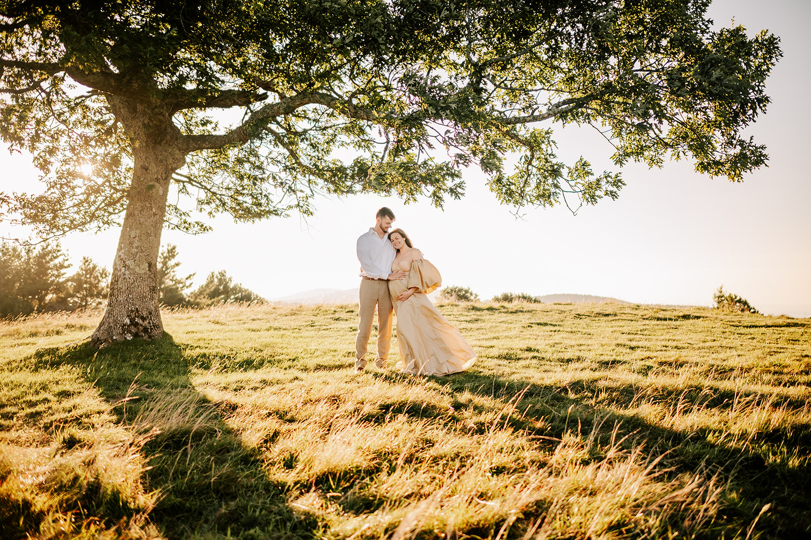 A happy expectant couple stand snuggling under a tree in a meadow at sunrise