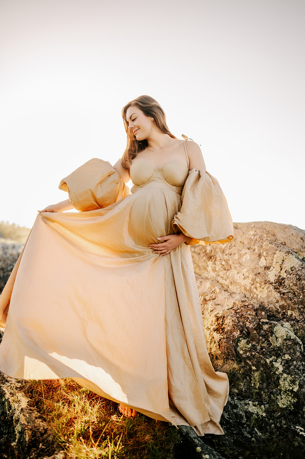 A expectant woman dances on a boulder in a gold maternity gown at sunrise