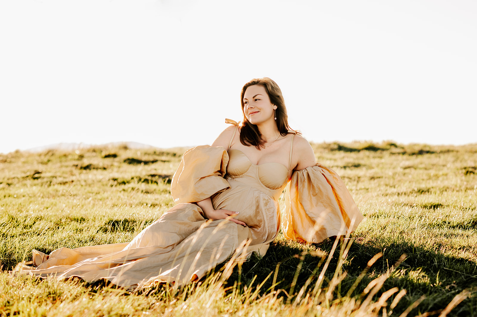 A happy mother to be in a gold maternity gown lays in a meadow at sunrise after finding prenatal chiropractors in Winston-Salem